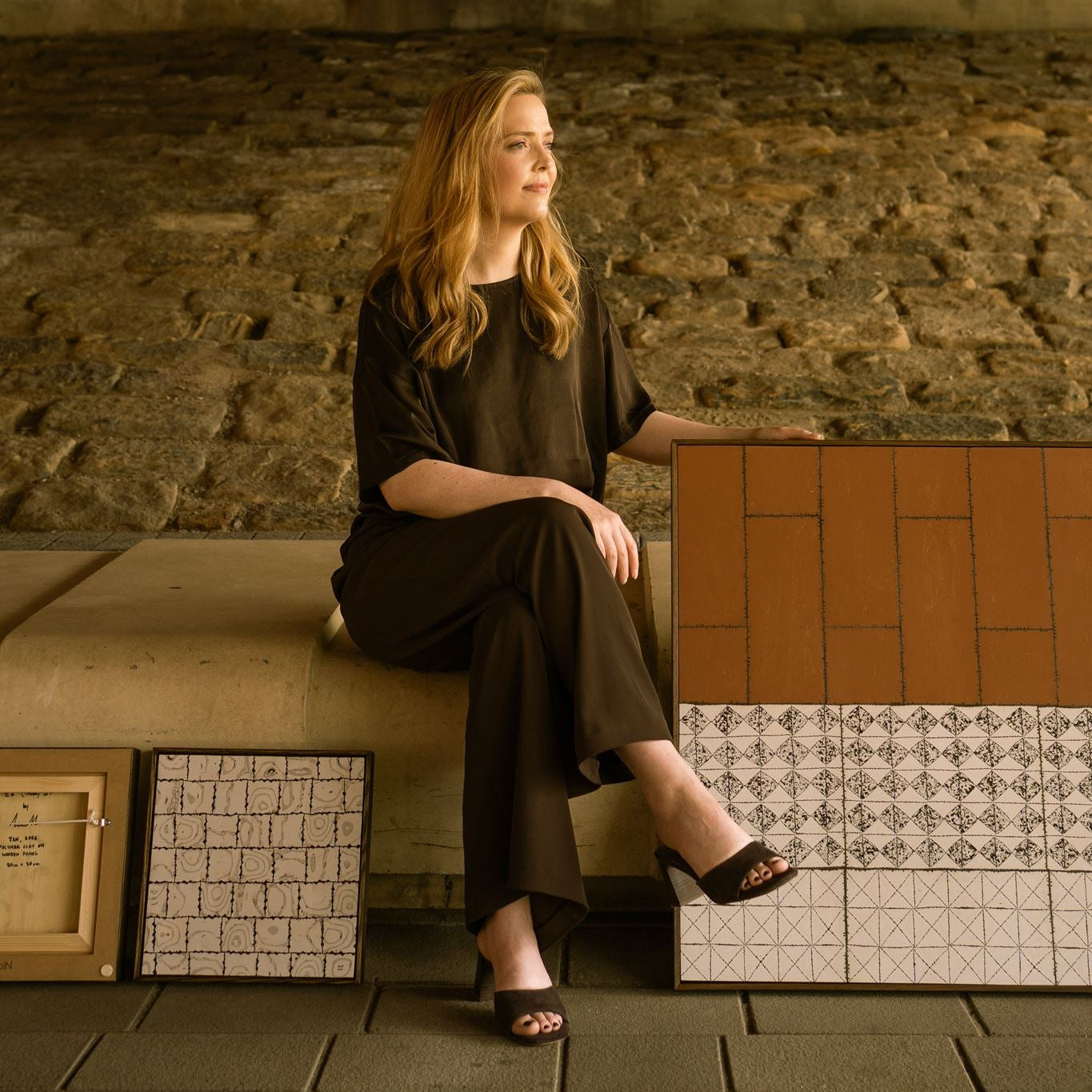 Anna Mills seated beside her contemporary abstract polymer clay artworks, photographed within a brutalist architectural setting of stone and concrete.