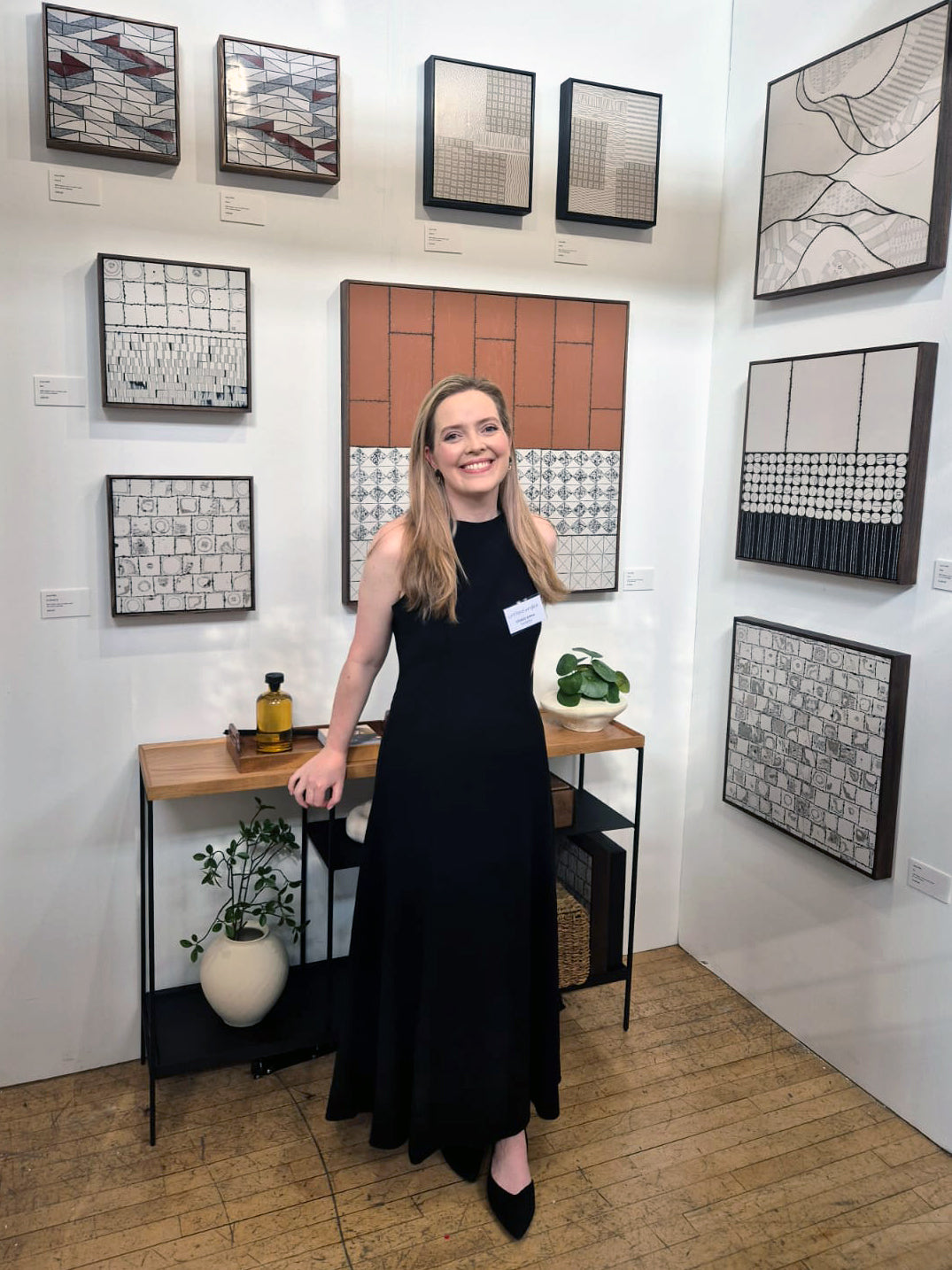 Anna Mills standing in her exhibition at Untitled Artists Fair, Chelsea Old Town Hall, London. She is wearing a long black dress and smiling in front of a display of framed abstract polymer clay artworks in muted black, white and terracotta tones. The exhibition stand features a simple oak console table with plants and carefully arranged decorative objects, reflecting the architectural and minimalist aesthetic of the collection.