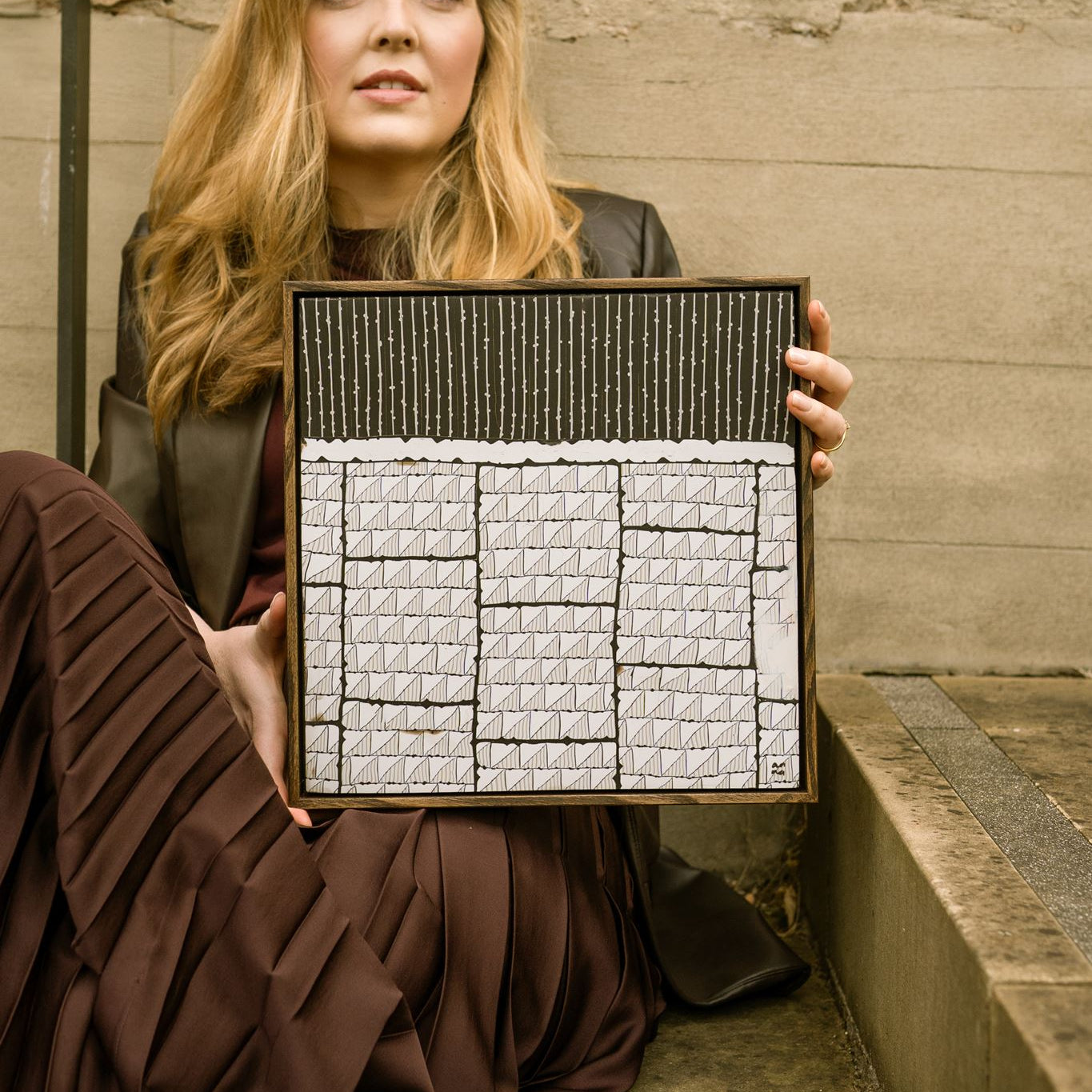 Anna Mills holding Steppe, a framed abstract dark brown and off-white polymer clay artwork featuring layered geometric patterns and linear textures.