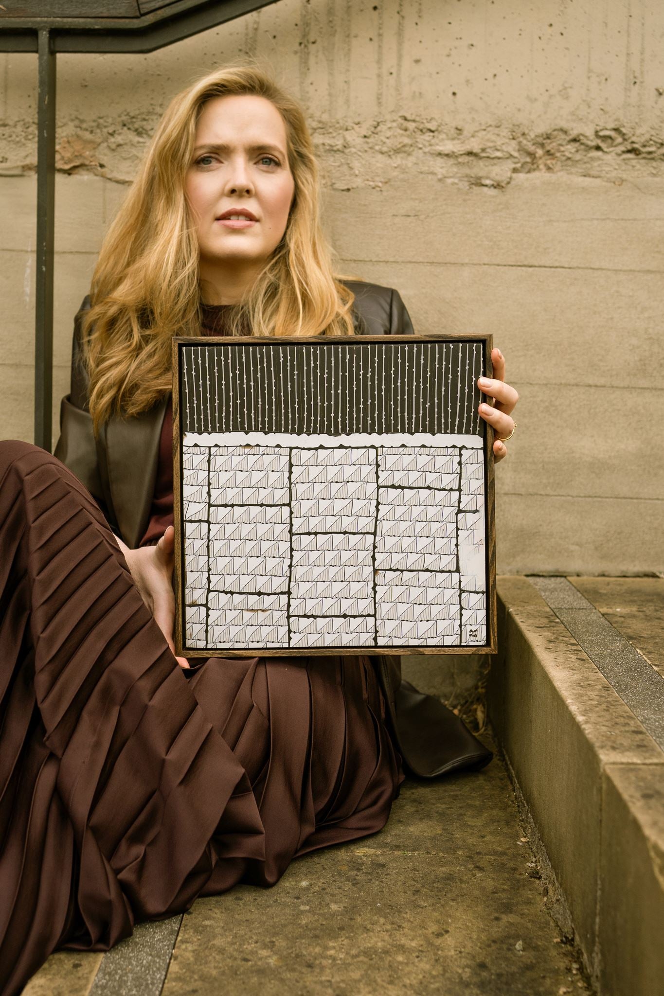 Anna Mills holding Steppe, a framed abstract dark brown and off-white polymer clay artwork featuring layered geometric patterns and linear textures.