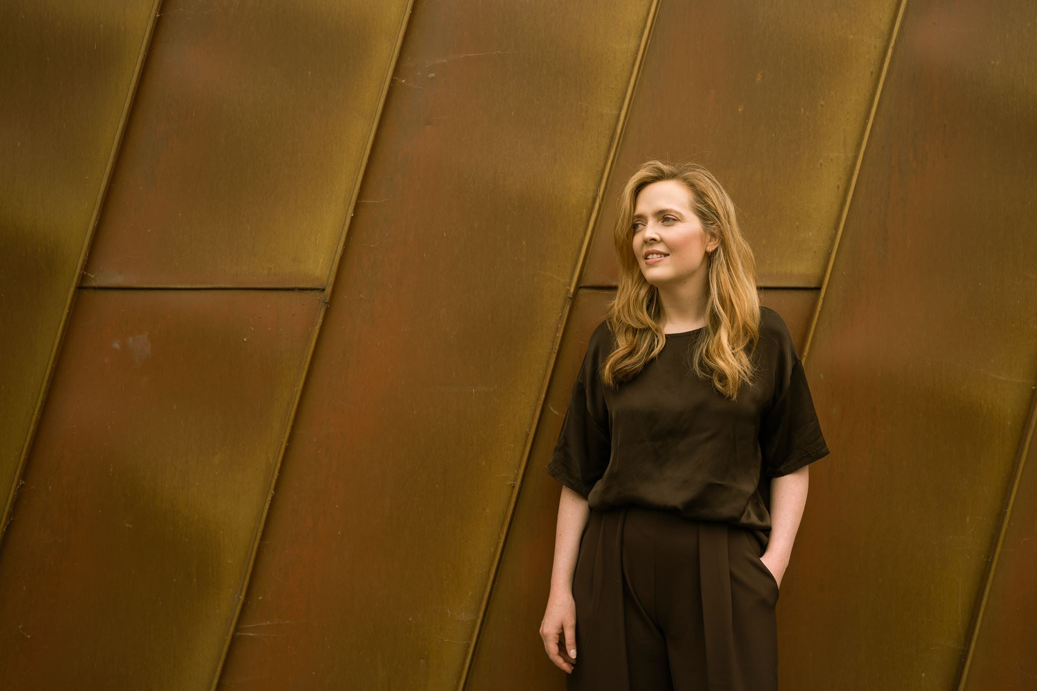 Portrait of visual artist Anna Mills standing against a weathered Corten steel wall with strong diagonal lines. She wears a relaxed dark brown top and tailored trousers, looking into the distance with one hand in her pocket. The warm rust tones of the architecture contrast with her minimalist clothing, reflecting the refined, material-led aesthetic of Studio Anna.