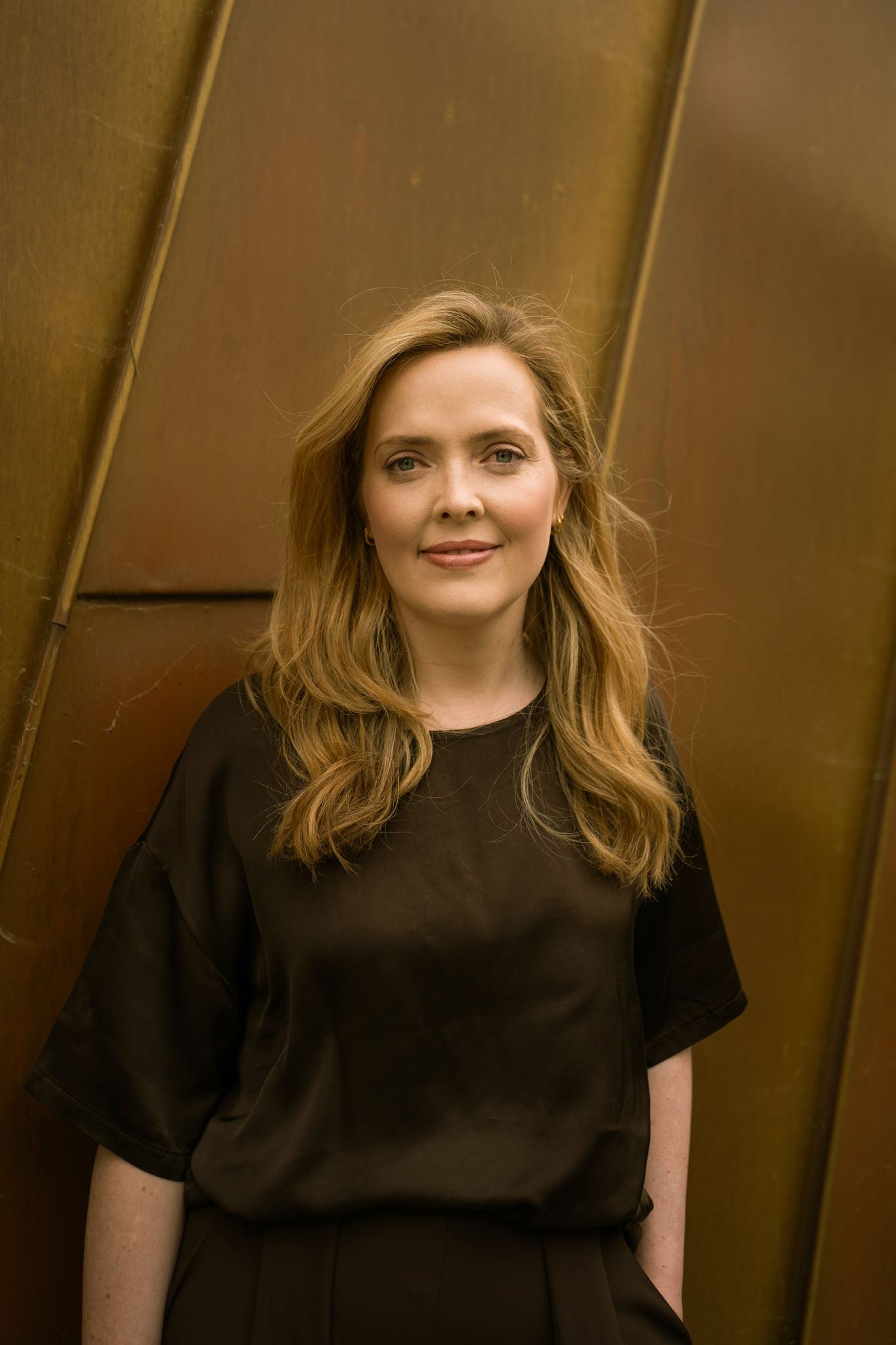 Close-up portrait of visual artist Anna Mills standing in front of a weathered Corten steel wall with bold diagonal lines. She wears a relaxed dark brown top and looks directly at the camera with a calm expression. The warm, earthy tones of the steel complement her minimalist style and reflect the material-led, architectural aesthetic of Studio Anna.