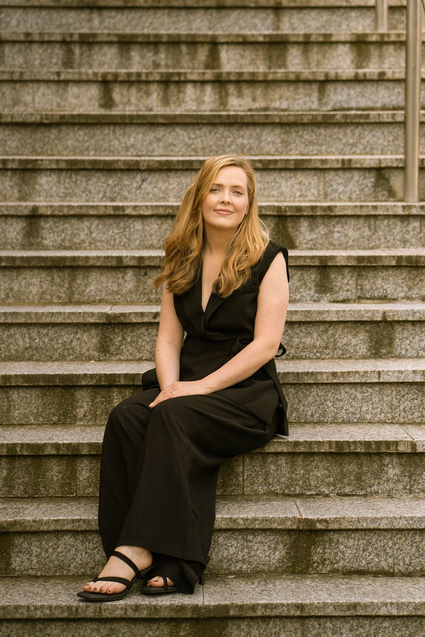 Portrait of visual artist Anna Mills seated on wide stone steps, wearing a sleeveless black wrap blazer, wide-leg trousers and black sandals. She faces the camera with a relaxed expression, framed by the strong horizontal lines and textured stone architecture that echo the refined, minimalist aesthetic of Studio Anna.