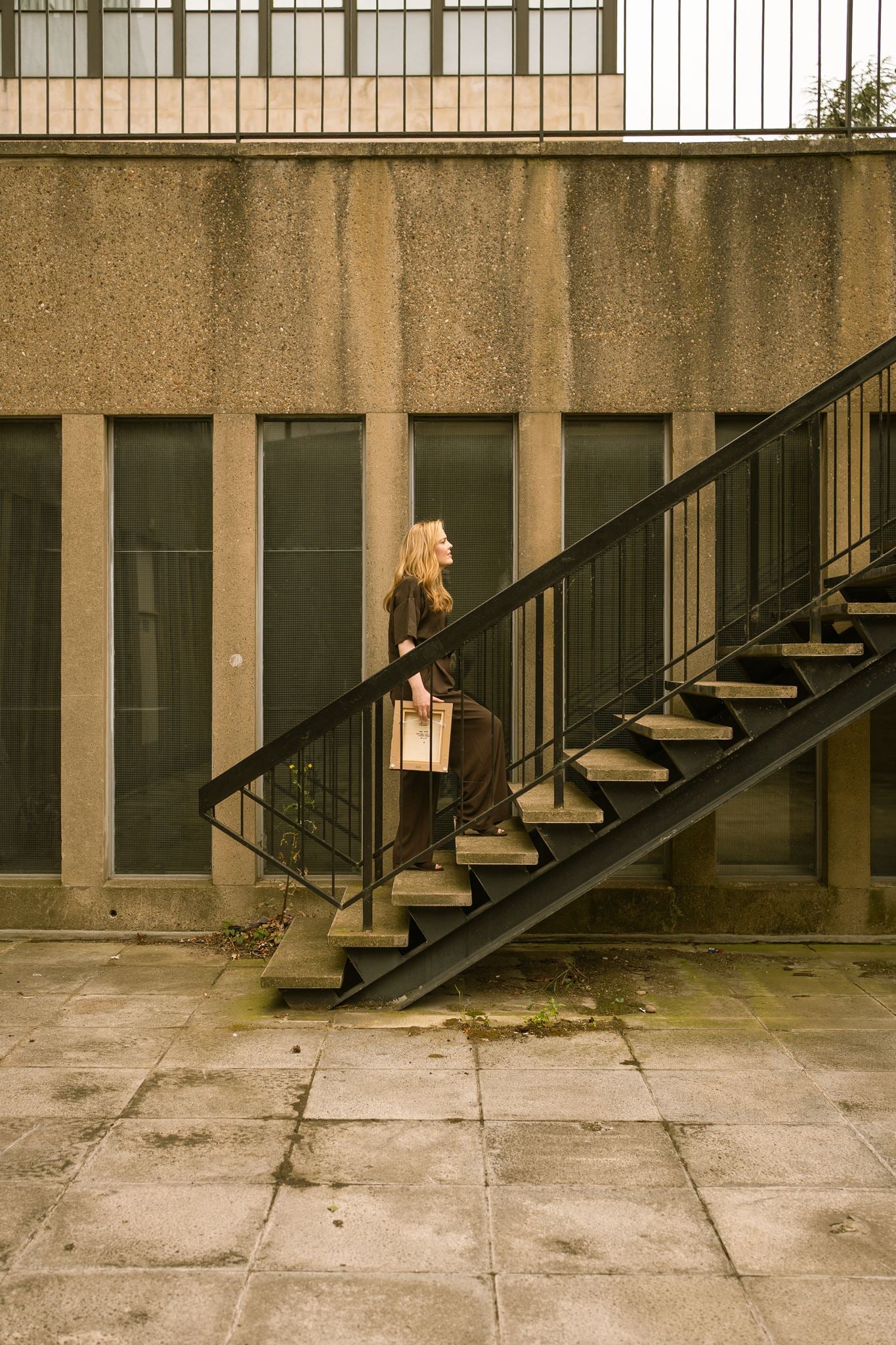 Artist Anna Mills carrying a framed artwork up an exterior concrete staircase against a Brutalist architectural backdrop.