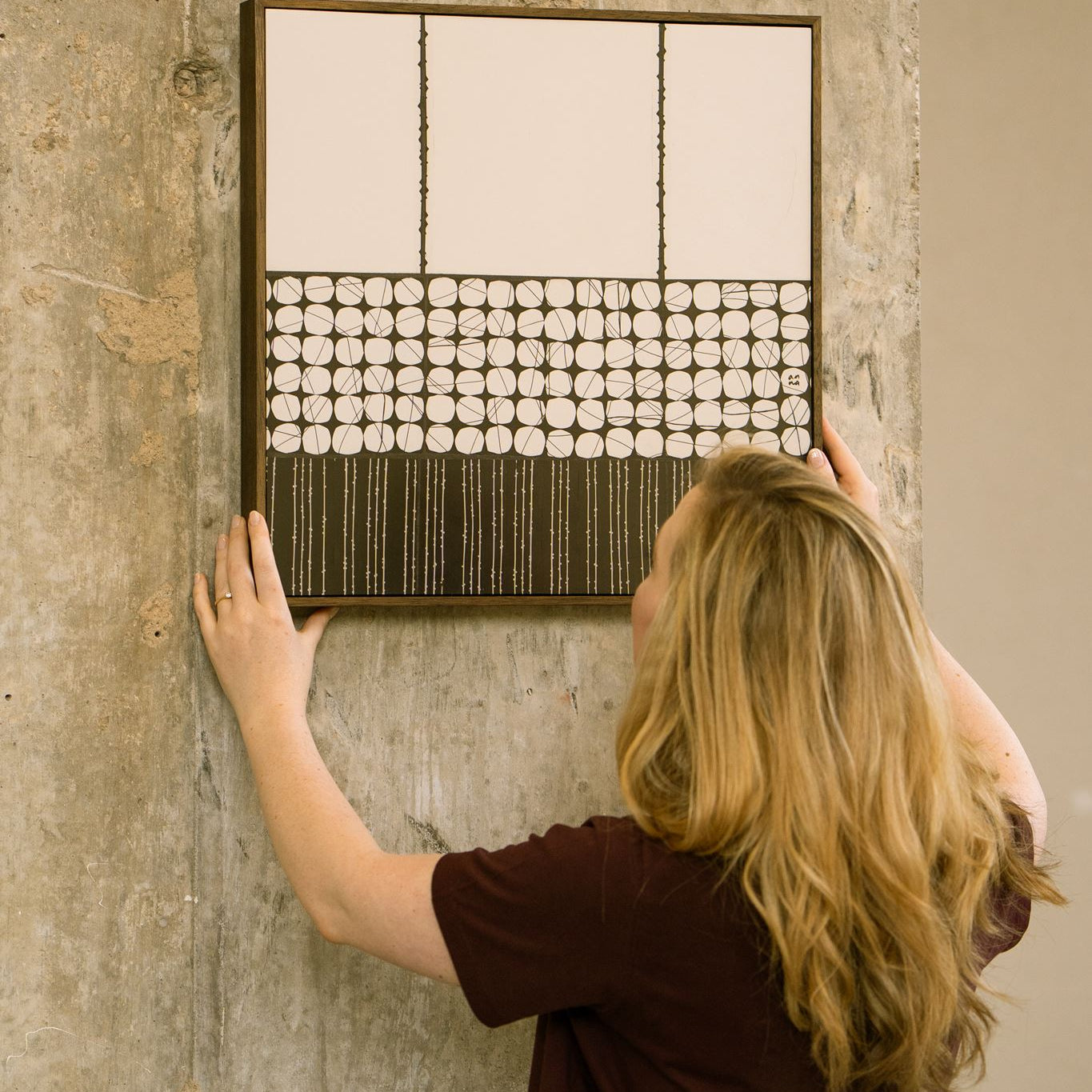 Artist Anna Mills positioning Heġe, a framed contemporary abstract polymer clay artwork with minimalist circular and striped geometric cane patterns, against a concrete wall.