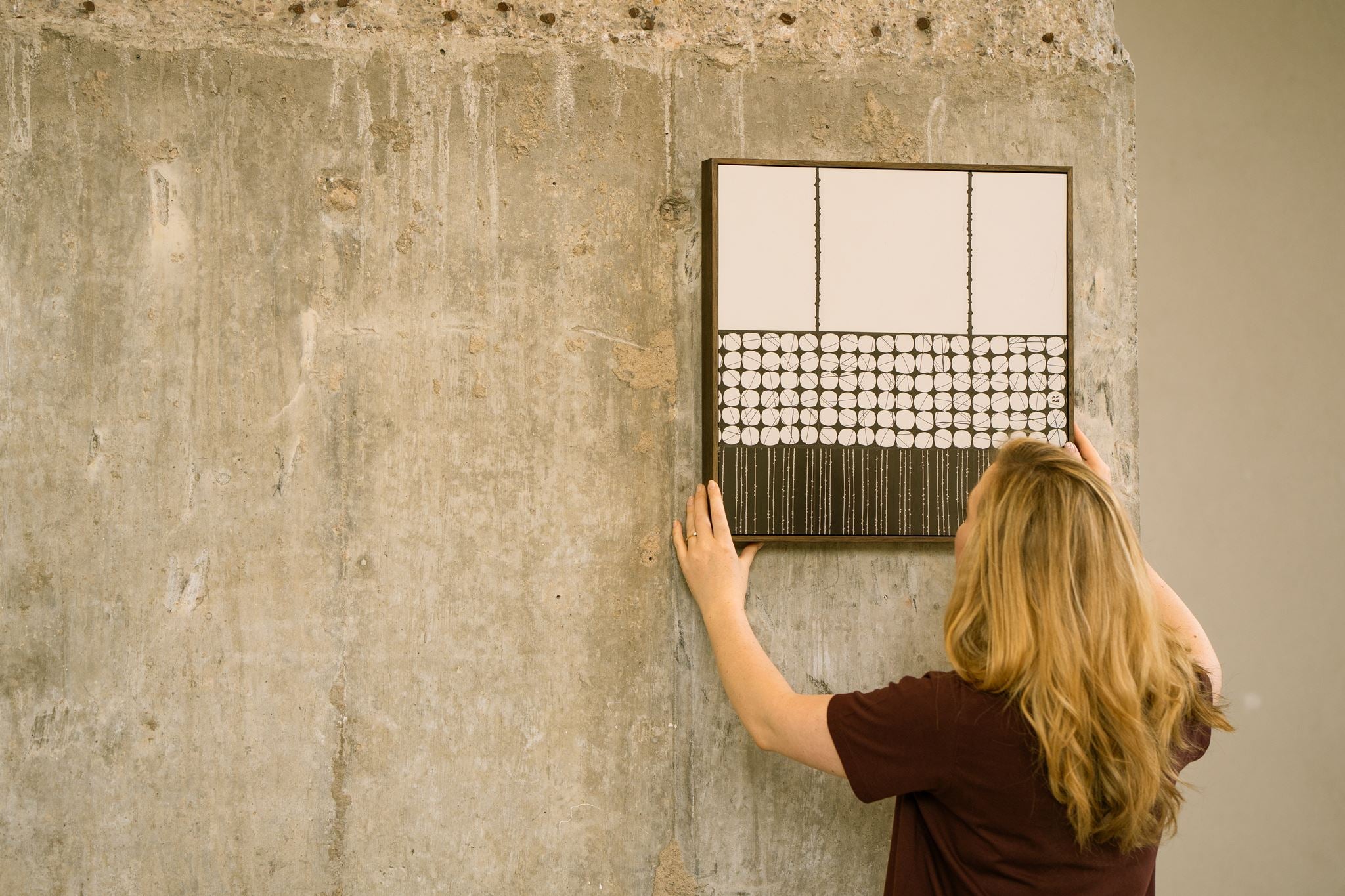 Artist Anna Mills positioning Heġe, a framed contemporary abstract polymer clay artwork with minimalist circular and striped geometric cane patterns, against a concrete wall.