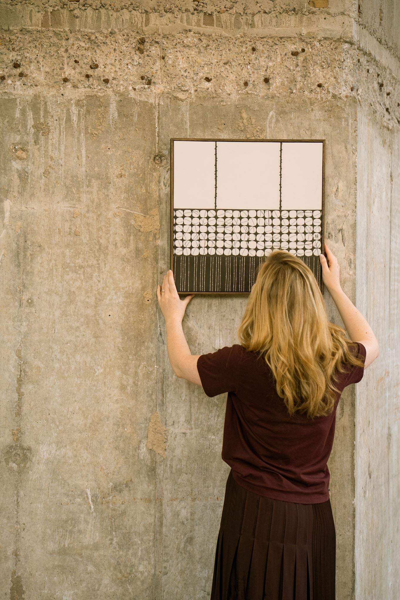 Artist Anna Mills positioning Heġe, a framed contemporary abstract polymer clay artwork with minimalist circular and striped geometric cane patterns, against a concrete wall.