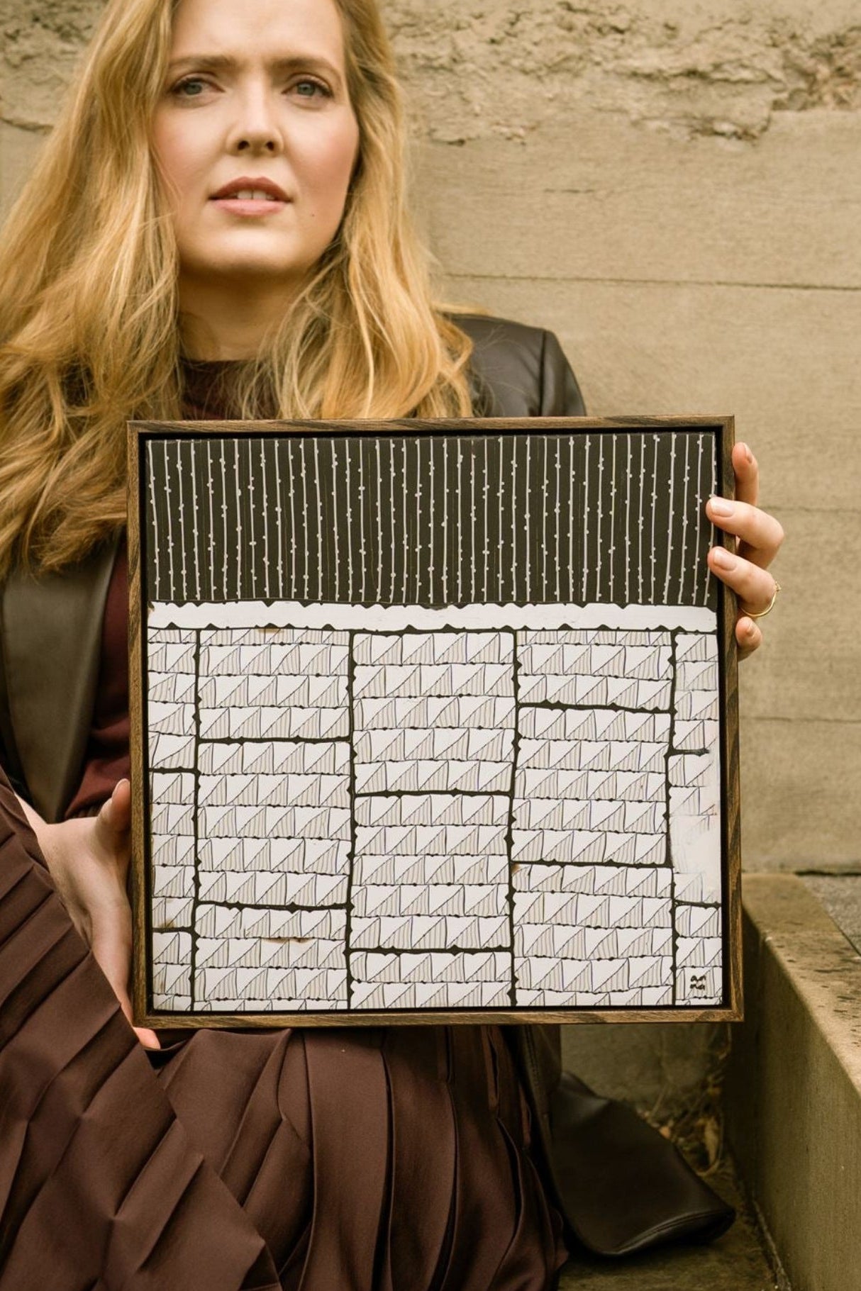 Portrait of visual artist Anna Mills seated on stone steps, holding 'Steppe', a framed contemporary polymer clay artwork in a dark English walnut wood veneer tray frame. The abstract composition features three columns of geometric black and white patterned forms beneath a band of fine vertical lines, photographed against a textured concrete wall in an urban setting.