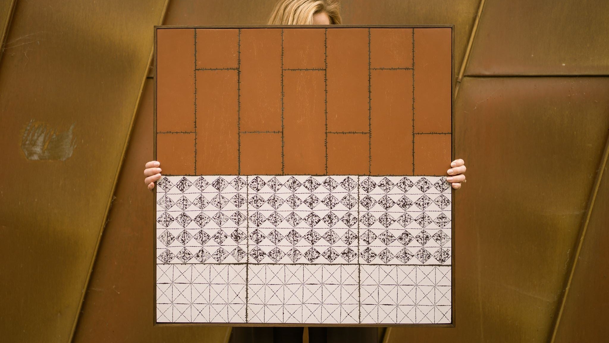 Visual artist Anna Mills stands behind her framed artwork, Drȳge, holding it in front of her so that only her hands and the top of her hair are visible. The artwork combines a warm rust-coloured upper section with geometric black-and-white patterned bands below, set against a weathered Corten steel architectural backdrop that complements its earthy palette.