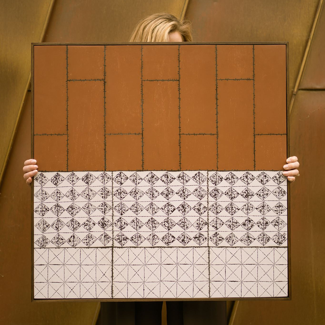 Visual artist Anna Mills stands behind her framed artwork, Drȳge, holding it in front of her so that only her hands and the top of her hair are visible. The artwork combines a warm rust-coloured upper section with geometric black-and-white patterned bands below, set against a weathered Corten steel architectural backdrop that complements its earthy palette.