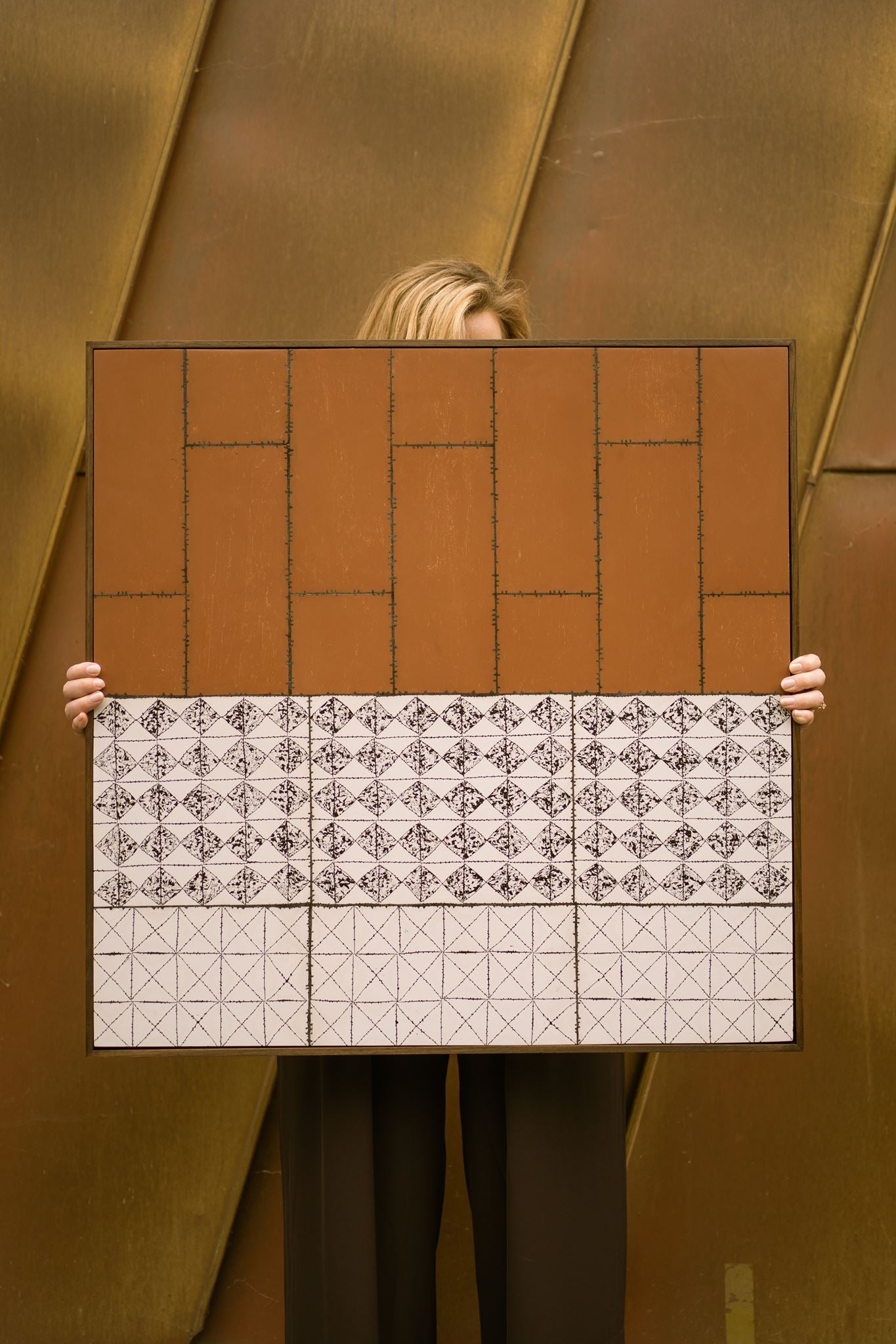 Visual artist Anna Mills stands behind her framed artwork, Drȳge, holding it in front of her so that only her hands and the top of her hair are visible. The artwork combines a warm rust-coloured upper section with geometric black-and-white patterned bands below, set against a weathered Corten steel architectural backdrop that complements its earthy palette.