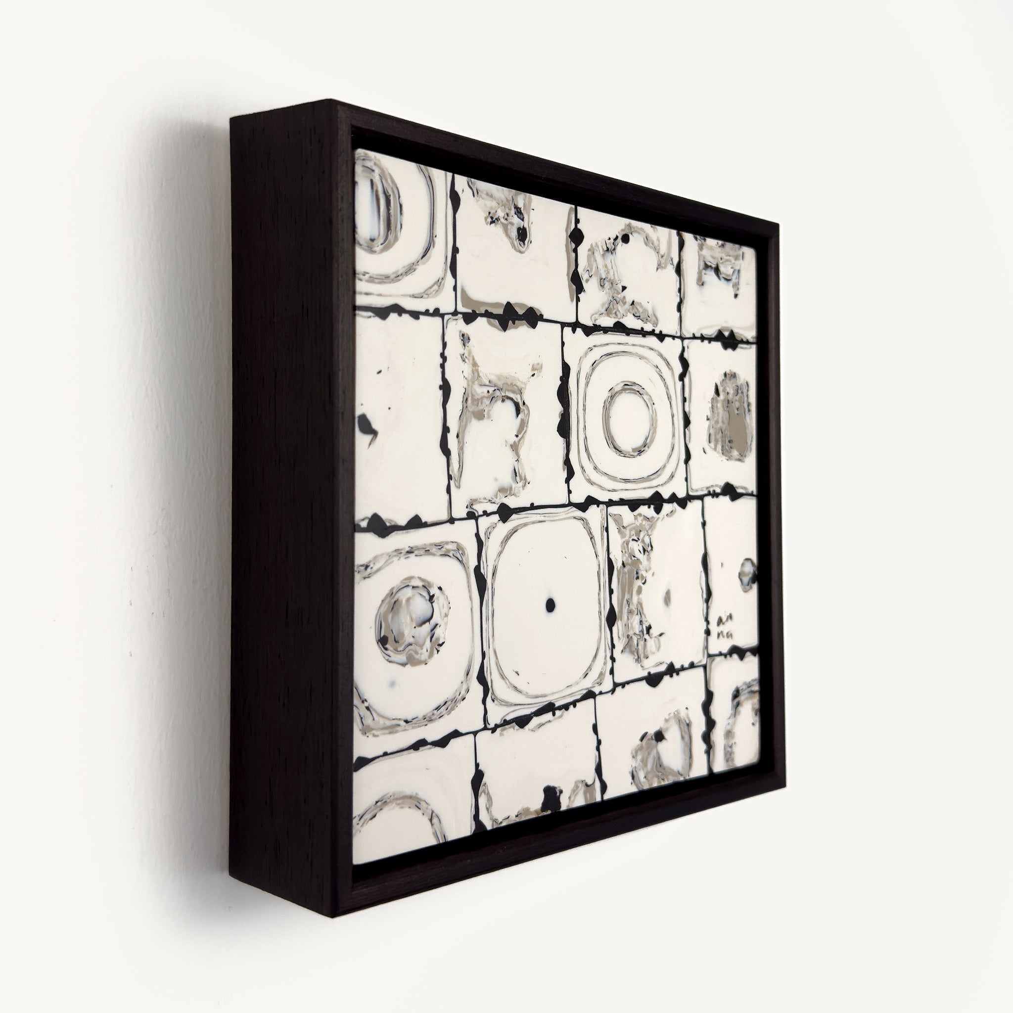 Ēa (Study III), a contemporary abstract polymer clay artwork composed of a grid of off-white toned square tiles featuring organic circular impressions, fragmented textures and subtle grey marbling. Photographed at an angle. Dark brown grout-like lines and irregular edges emphasise the handmade surface, creating a quiet dialogue between order and erosion. Presented in a solid wood veneer tray frame finished in dark English walnut, measuring 17 × 17 cm.