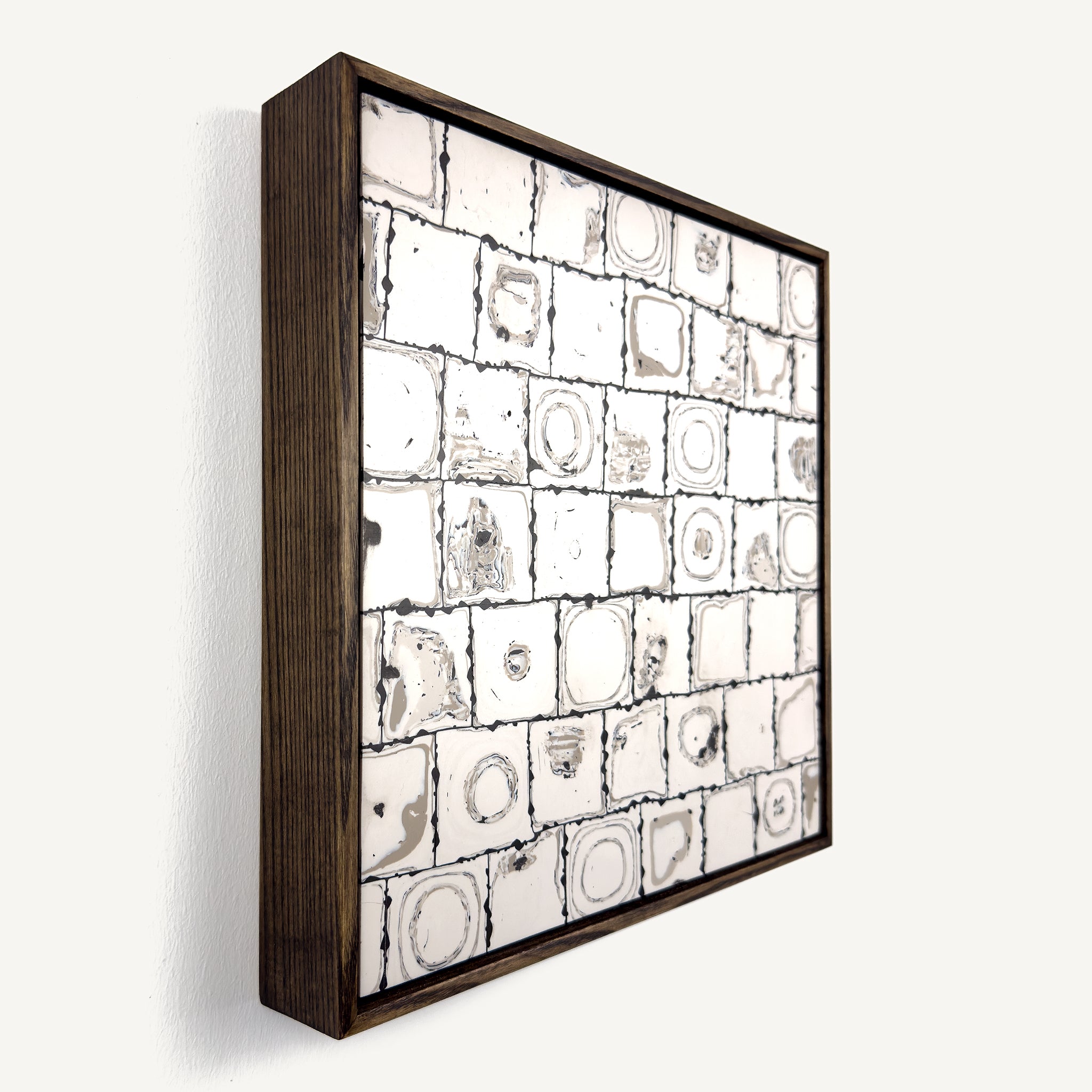 Ēa (Study II), a contemporary abstract polymer clay artwork, by Anna Mills, composed of a grid of individually hand-cut cane tiles, each revealing unique concentric patterns created from layered clay. Photographed at an angle. Soft ivory, warm grey and dark brown/black tones evoke geological formations and topographic contours, while the irregular tile edges and subtle surface variations celebrate the handmade process. Presented in a solid wood tray frame finished in dark English walnut.