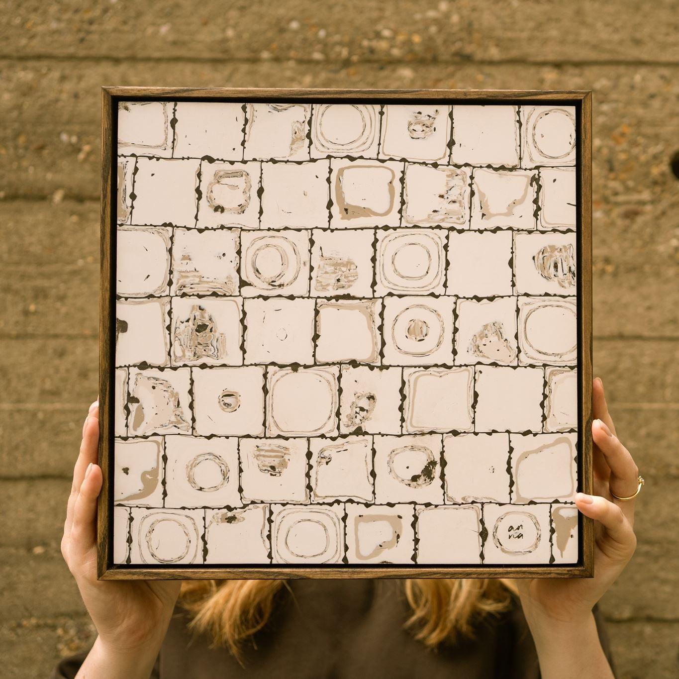 Artist Anna Mills holding up Ēa (Study II), a contemporary abstract polymer clay artwork featuring hand-cut square cane slices arranged in a geometric grid with layered off-white and dark brown patterns the resemble ripples in water.