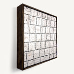 Ēa (Study I), a contemporary abstract polymer clay artwork, by Anna Mills, composed of a grid of individually hand-cut cane tiles, each revealing unique concentric patterns created from layered clay. Photographed at an angle. Soft ivory, warm grey and dark brown/black tones evoke geological formations and topographic contours, while the irregular tile edges and subtle surface variations celebrate the handmade process. Presented in a solid wood tray frame finished in dark English walnut.