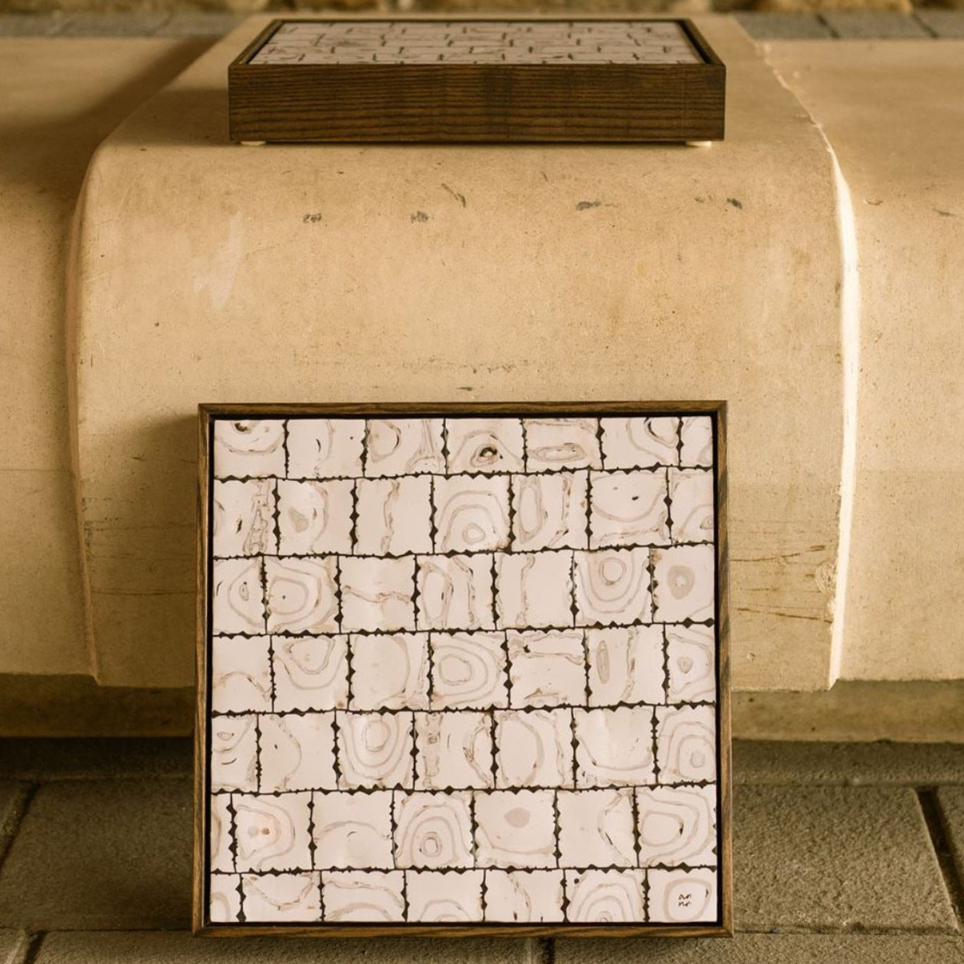 Framed abstract polymer clay artwork, titled Ēa (Study I), composed of hand-cut square tiles with layered off-white and dark brown organic patterns, displayed against a warm stone backdrop.