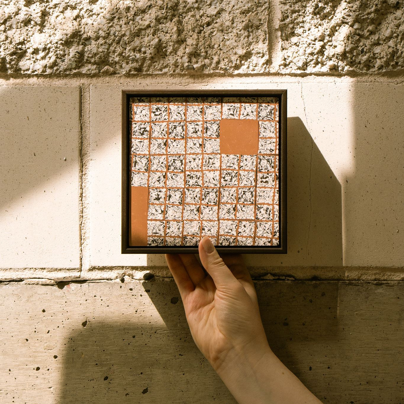 Greót II, framed contemporary abstract polymer clay artwork with textured black, white and terracotta square cane slices, shown held against a concrete wall in natural light.