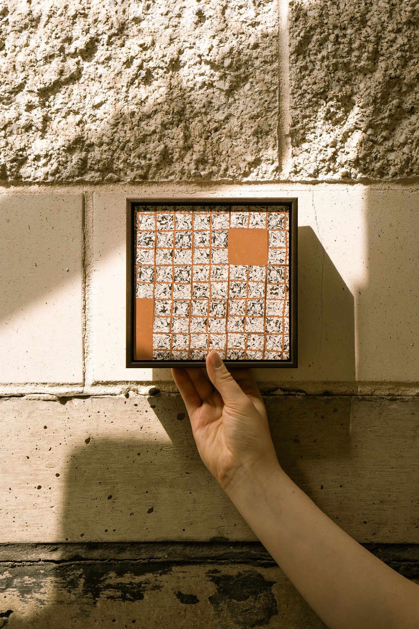 Greót II, framed contemporary abstract polymer clay artwork with textured black, white and terracotta square cane slices, shown held against a concrete wall in natural light.