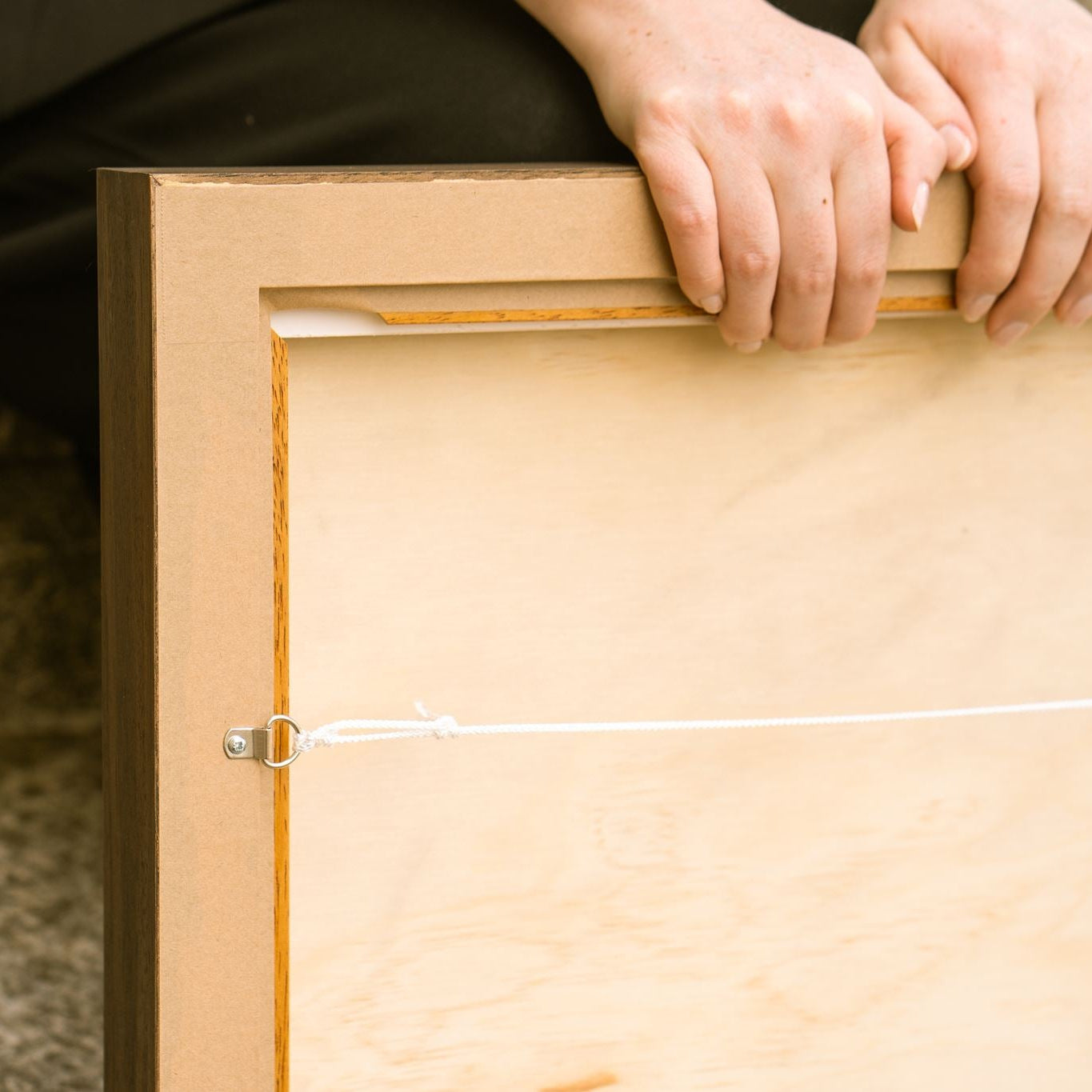 Close-up of the reverse corner of a professionally framed Studio Anna artwork, showing the walnut-stained tray frame, wooden backing board, D-ring hanging hardware and white hanging cord. The artist's hands rest on the frame, highlighting the craftsmanship, quality materials and ready-to-hang finish.