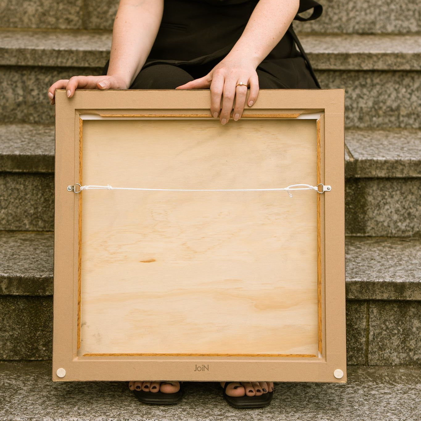 Reverse of a professionally framed Studio Anna artwork, showing the wooden backing board, D-ring hanging hardware and white hanging cord. The artist holds the frame while seated on stone steps, demonstrating the clean finish and ready-to-hang presentation of the artwork.