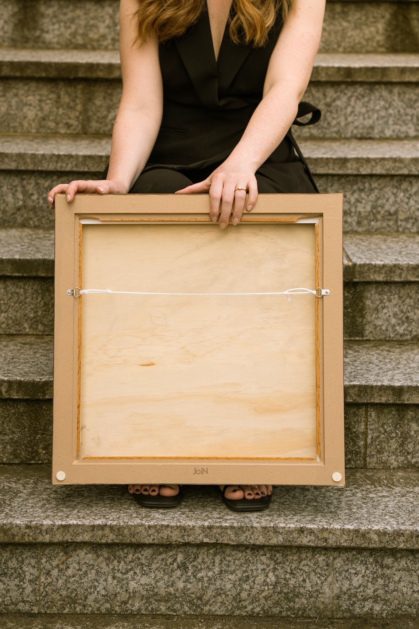 Reverse of a professionally framed Studio Anna artwork, showing the wooden backing board, D-ring hanging hardware and white hanging cord. The artist holds the frame while seated on stone steps, demonstrating the clean finish and ready-to-hang presentation of the artwork.